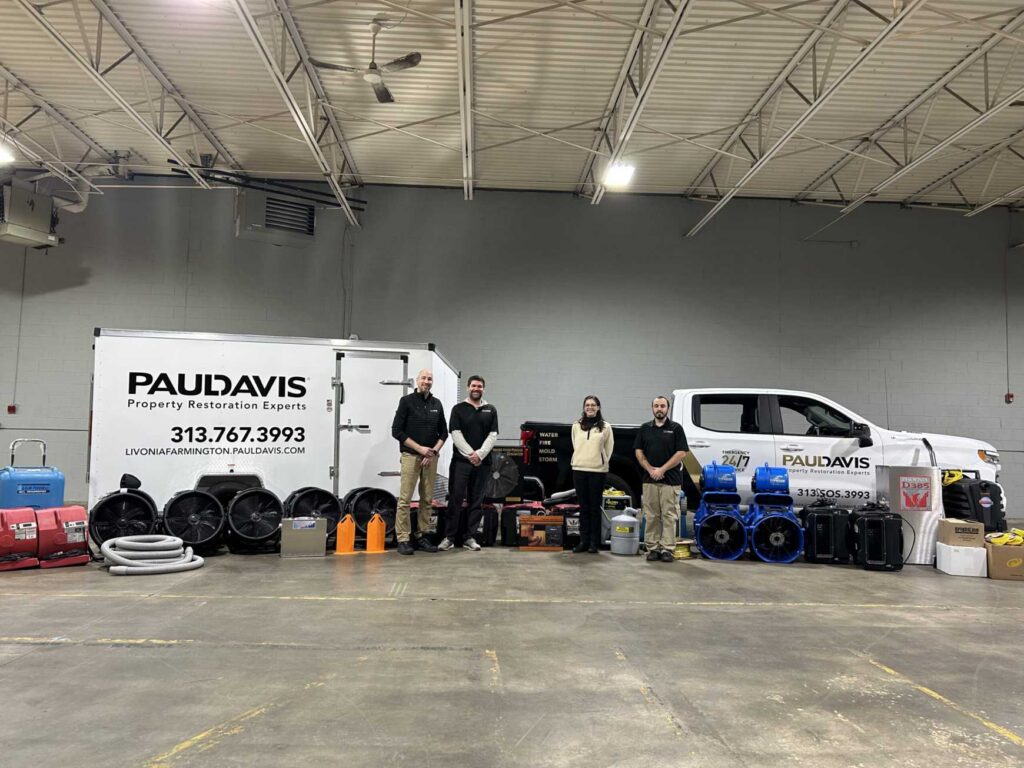 Paul Davis Restoration of Livonia/Farmington, MI team standing inside a warehouse with a service trailer, company truck, and a wide range of water, fire, and mold restoration equipment including air movers, dehumidifiers, and hoses.